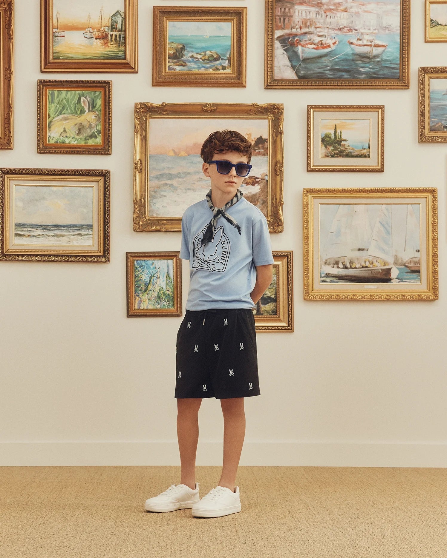 A child wearing sunglasses, the Psycho Bunny Kids Melbourne Graphic Tee - B0U410F200 in light blue made from eco-friendly Peruvian Pima cotton, black shorts, and white sneakers stands in front of a wall with framed landscape paintings.