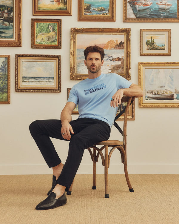 A man sits on a wooden chair wearing a light blue Psycho Bunny MENS BERNI EMBROIDERED WORDMARK GRAPHIC TEE made in Peru, paired with black pants and shoes. Behind him, framed landscape and seascape paintings adorn a white wall, while a beige carpet adds soft tones to the scene.