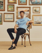 A man sits on a wooden chair wearing a light blue Psycho Bunny MENS BERNI EMBROIDERED WORDMARK GRAPHIC TEE made in Peru, paired with black pants and shoes. Behind him, framed landscape and seascape paintings adorn a white wall, while a beige carpet adds soft tones to the scene.
