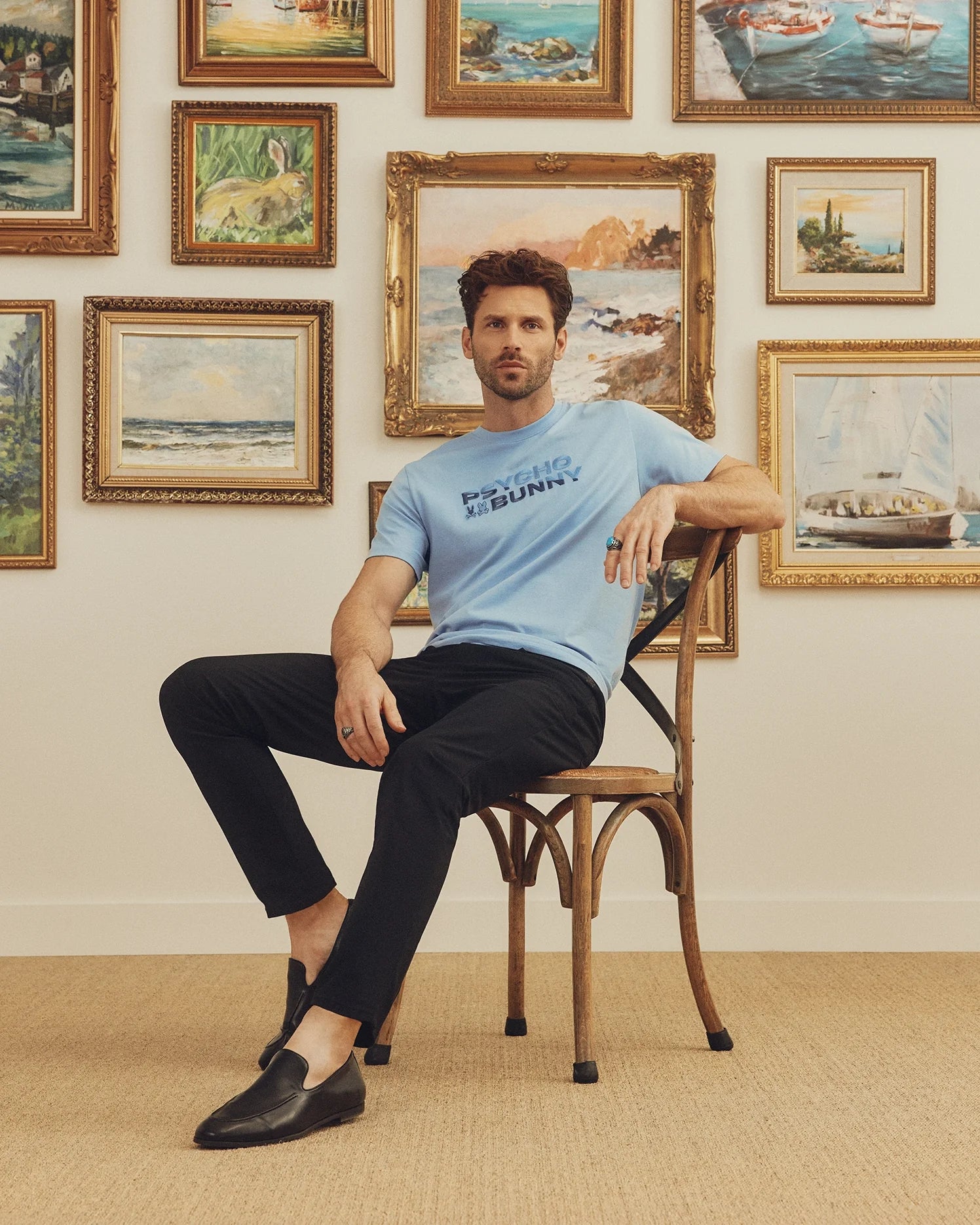 A man sits on a wooden chair wearing a light blue Psycho Bunny MENS BERNI EMBROIDERED WORDMARK GRAPHIC TEE made in Peru, paired with black pants and shoes. Behind him, framed landscape and seascape paintings adorn a white wall, while a beige carpet adds soft tones to the scene.