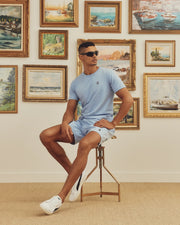 A man wearing a light blue Psycho Bunny MENS CLASSIC CREW NECK TEE and shorts sits on a stool with sunglasses. Behind him, a wall displays vibrant landscape paintings. He wears white sneakers.