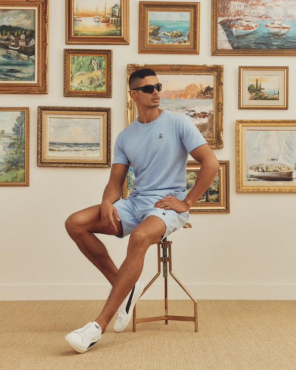 A man wearing a light blue Psycho Bunny MENS CLASSIC CREW NECK TEE and shorts sits on a stool with sunglasses. Behind him, a wall displays vibrant landscape paintings. He wears white sneakers.