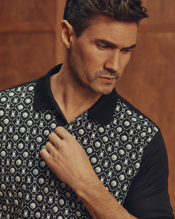 A man gazes downward, showcasing the Psycho Bunny MENS BECKETT PIQUE POLO (MT0100022), crafted from premium Pima cotton with a black collar and a geometric circle-floral design. The scene is set against a warm wooden backdrop.