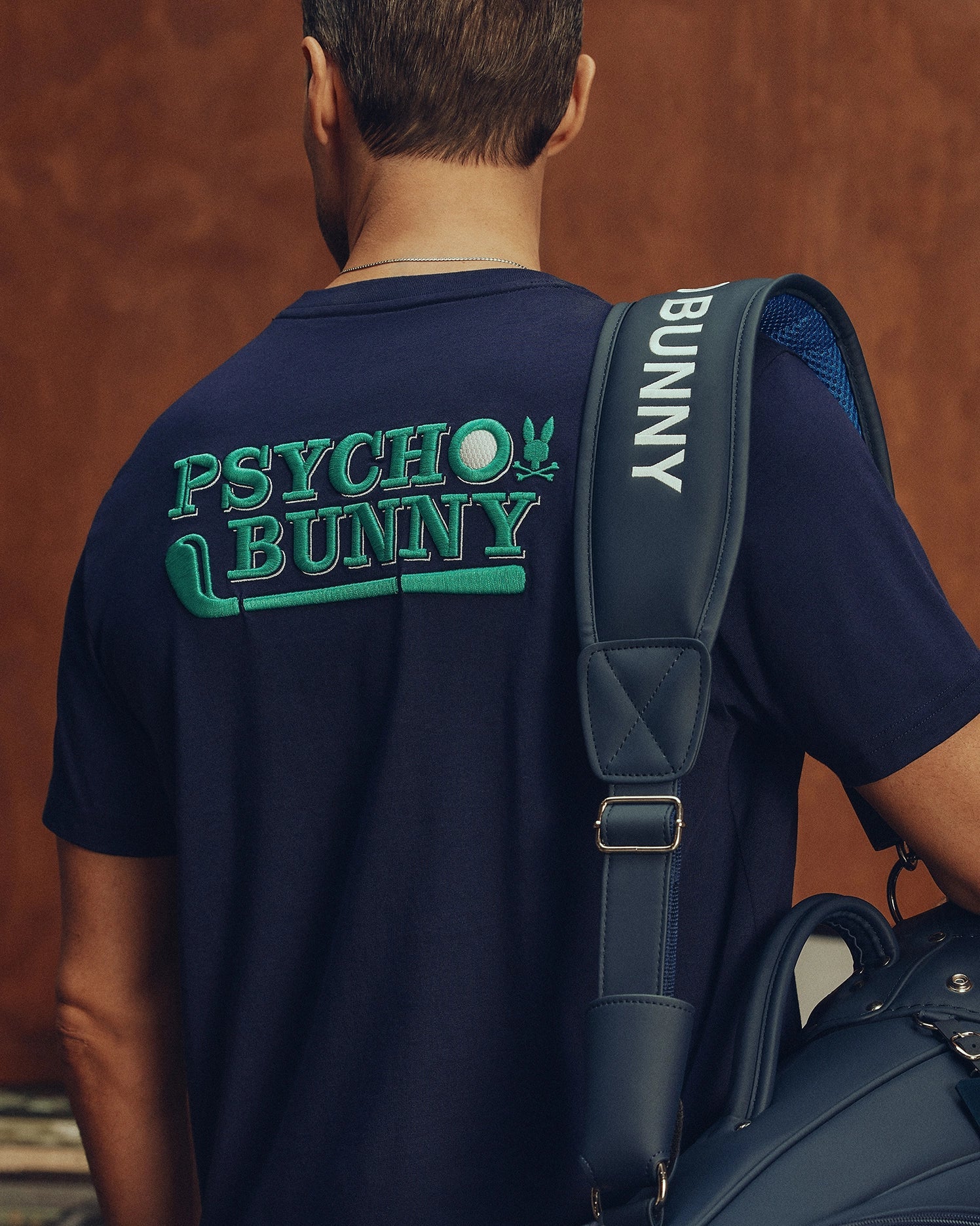 A person is seen from behind wearing a navy Psycho Bunny MENS DENIS GRAPHIC TEE - MT0200015 with "Psycho Bunny" in bold green on the back. A matching navy bag with an embroidered logo and "Bunny" on the strap rests on their shoulder against a subtle golf-inspired brownish wall.