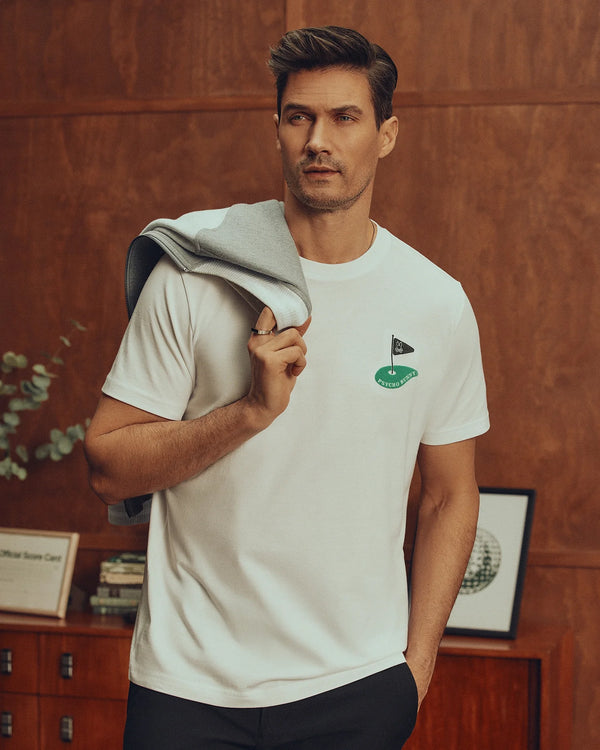 A man stands indoors wearing a white Psycho Bunny MENS DALE GRAPHIC TEE - MT0200016 with a green flag design and embroidered logo, holding a gray jacket over his shoulder. He has short hair and looks sideways, with a wooden cabinet and greenery behind him.