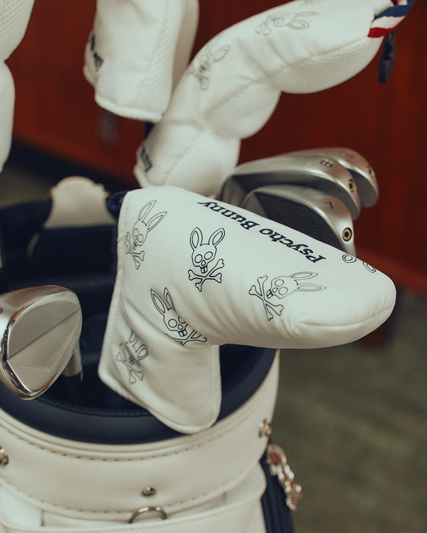 A golf bag stands indoors, showcasing putter covers adorned with a skull and crossbones design with bunny ears. Each white cover, marked "Psycho Bunny," adds edgy style to any golfer's collection. Enjoy the distinction of the MULTI BUNNY BLADE PUTTER COVER - PBMG4SH5 by Psycho Bunny.