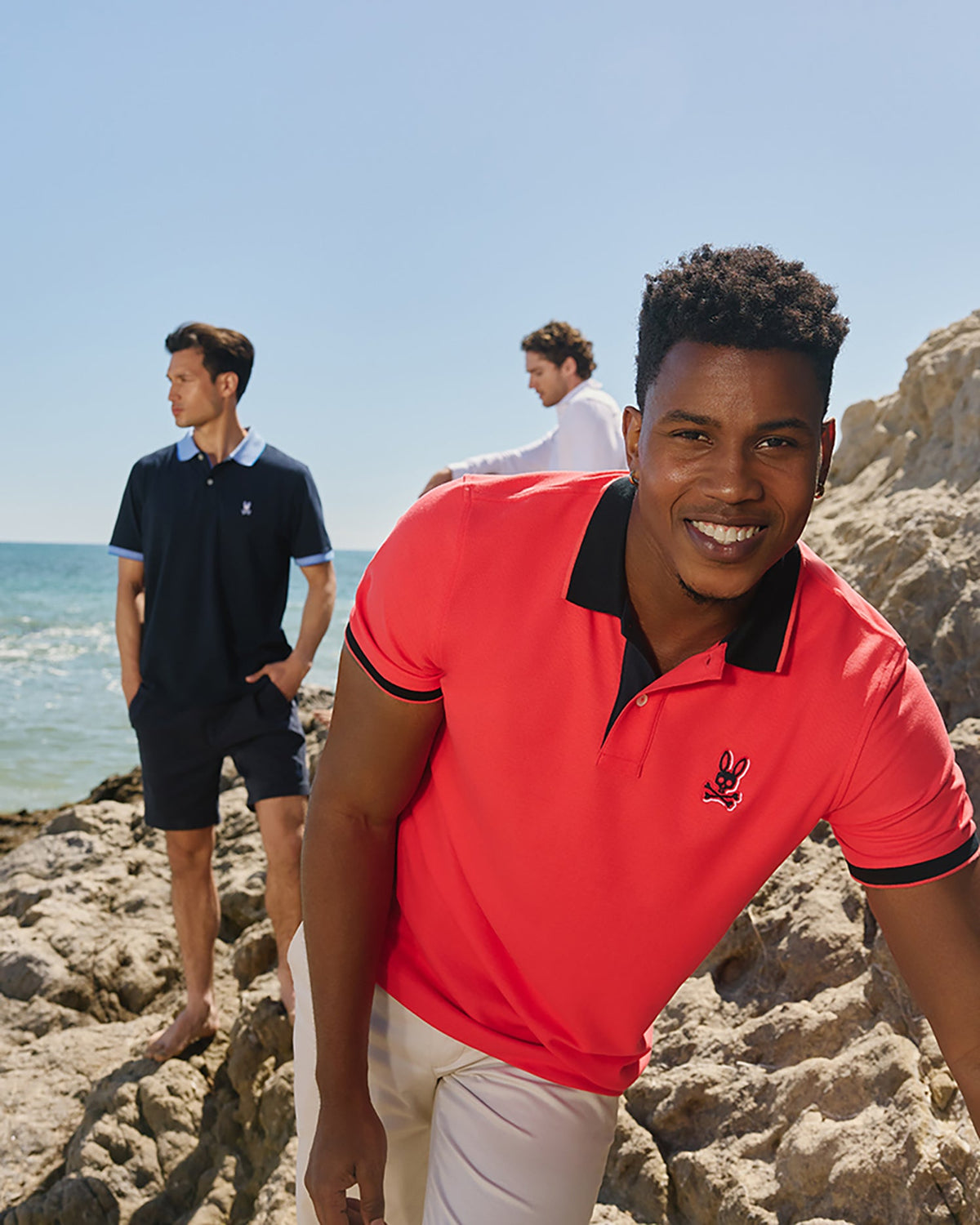 Three men are wearing polo shirts near a beach, with the ocean in the background. The man in the foreground wears a bright coral Psycho Bunny MENS KAYDEN PIQUE POLO - B6K675C200 and smiles at the camera. Another man in a navy polo with contrasting stripes looks into the distance, and the third stands further back in a white polo.
