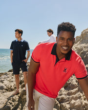 Three men are wearing polo shirts near a beach, with the ocean in the background. The man in the foreground wears a bright coral Psycho Bunny MENS KAYDEN PIQUE POLO - B6K675C200 and smiles at the camera. Another man in a navy polo with contrasting stripes looks into the distance, and the third stands further back in a white polo.