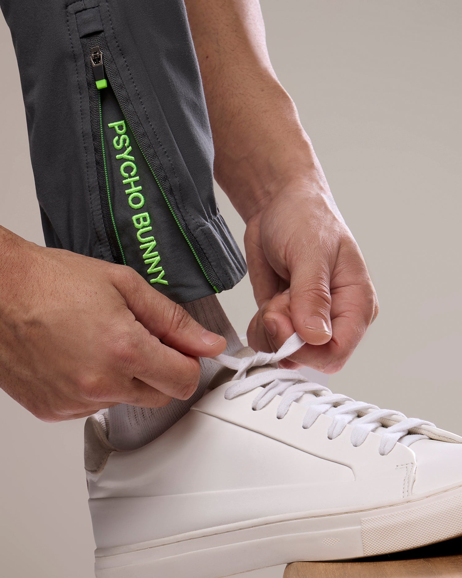 A person tying the laces of a white sneaker, wearing gray Psycho Bunny Men's Madison Commuter Pants with a green "psycho bunny" logo on the zipper. The focus is on the hands and shoe.