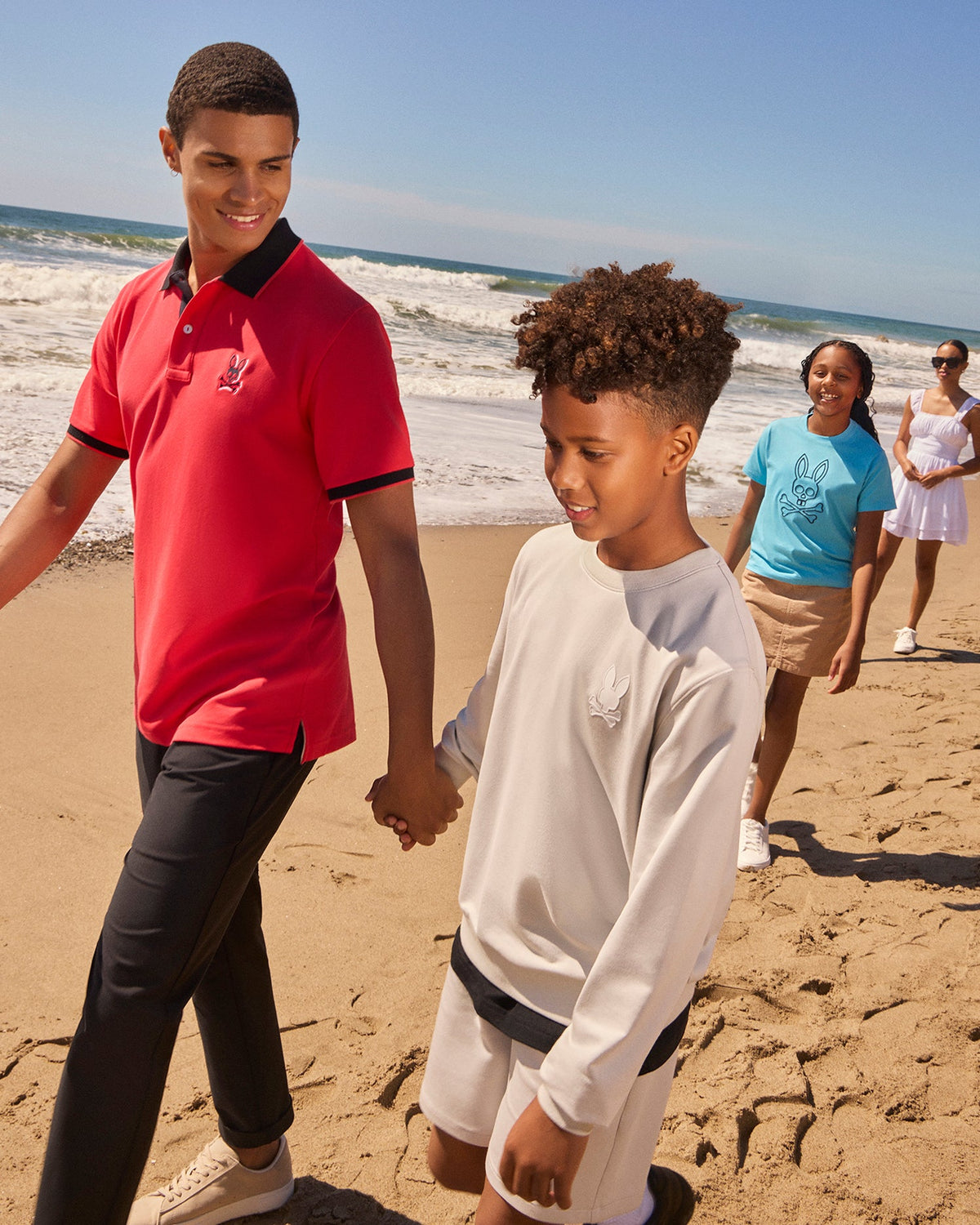 A group of four people, including two adults and two children, walk hand-in-hand along a beach. The adults wear casual beachwear—one in a red Psycho Bunny MENS KAYDEN PIQUE POLO - B6K675C200 and the other in a white dress with contrasting stripes—while the children wear comfortable clothing, smiling and enjoying the sunny weather.