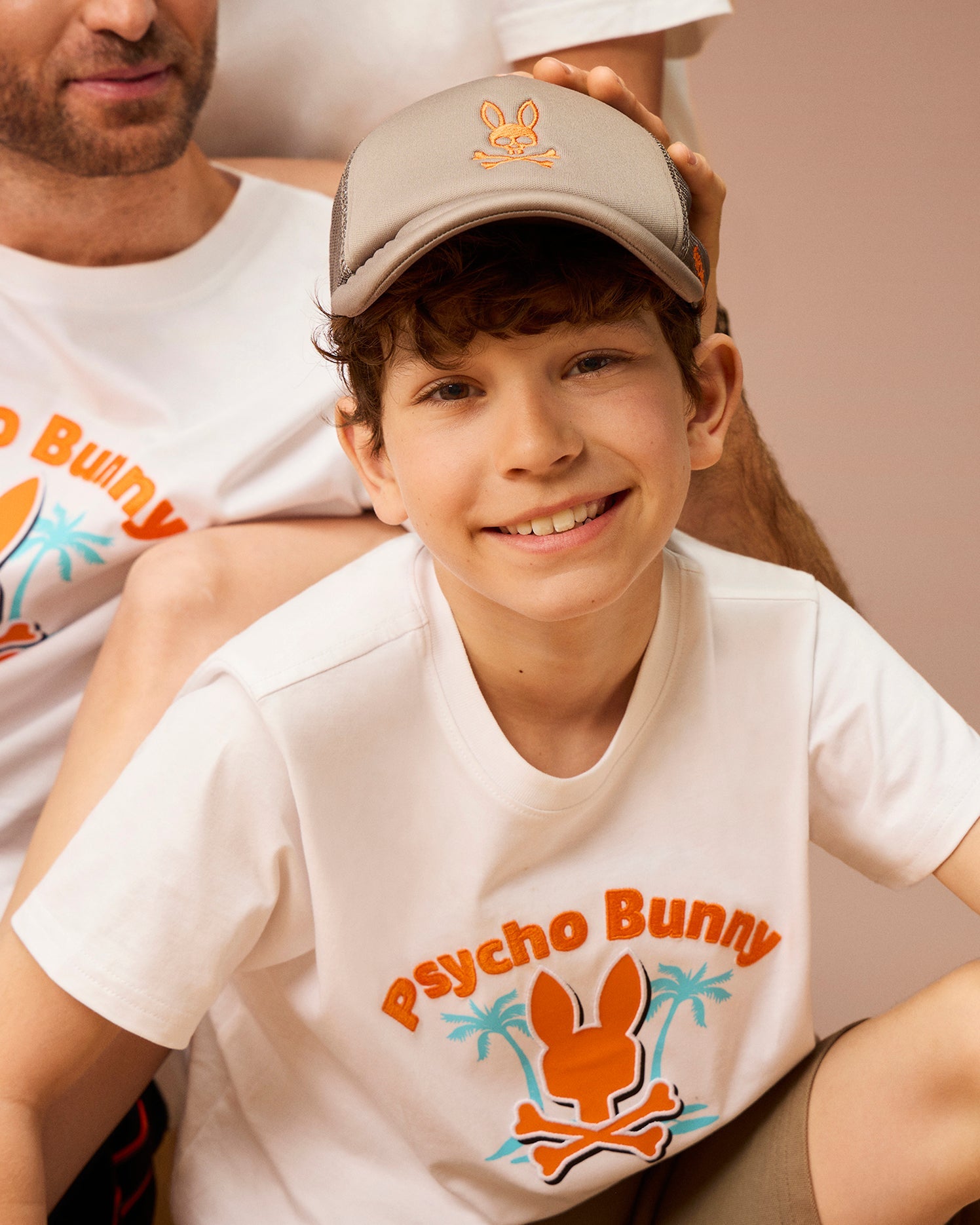 A smiling boy in a KIDS PARIS TRUCKER CAP - B0A591C200 by Psycho Bunny and a white t-shirt featuring an orange Bunny logo and palm trees sits in front of a man. They both wear matching shirts, with the man's arm resting on the boy's shoulder, creating a charming, coordinated look.