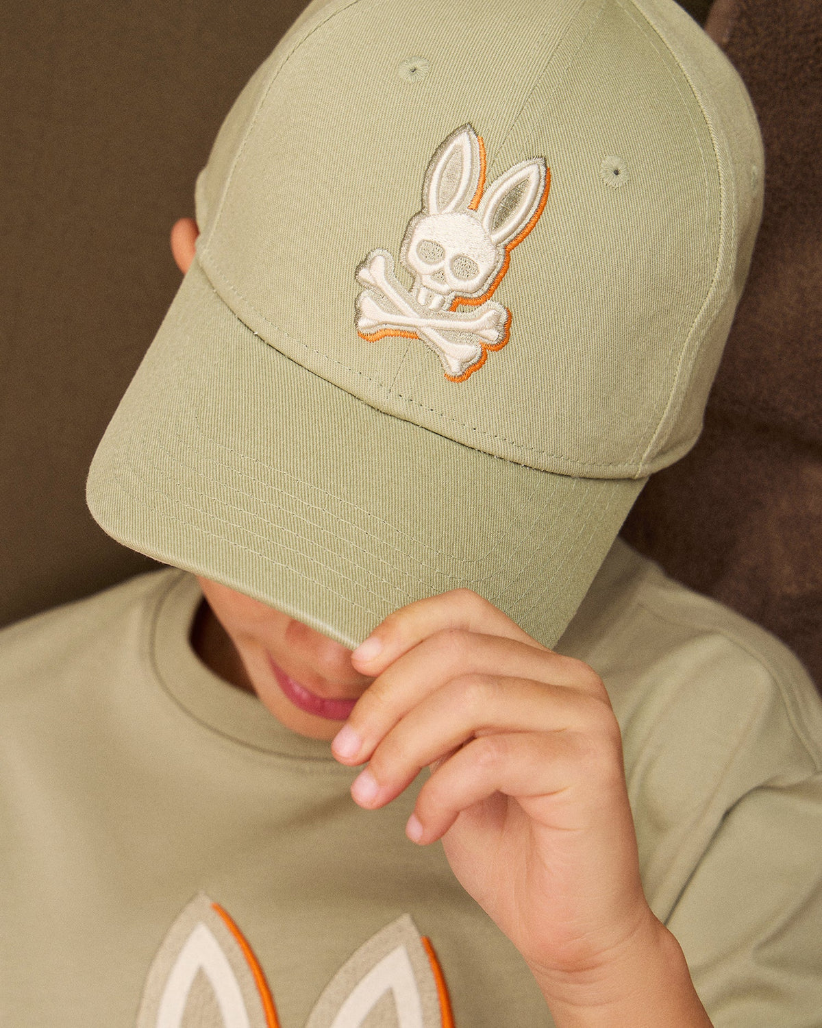 Close-up of a person wearing a light green KIDS KAYDEN BASEBALL CAP - B0A677C200 and shirt with matching rabbit and skull designs. The person tilts their head slightly downward, a hand adjusting the cap's brim adorned with an embroidered Psycho Bunny logo, partially obscuring their face.