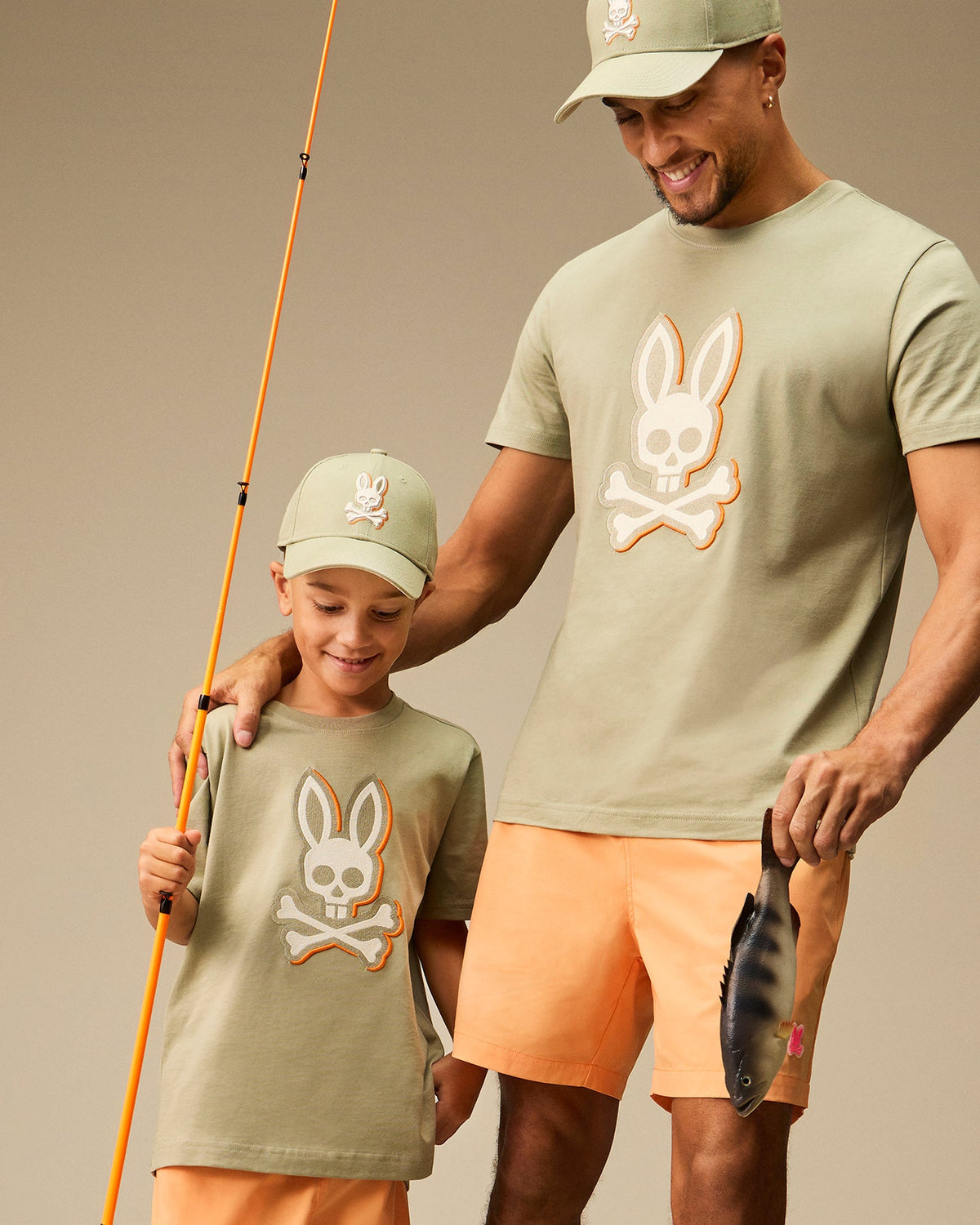 A man and a young boy, possibly father and son, are seen smiling and dressed in matching outfits with an embroidered Bunny logo. Both are wearing sage green cotton twill baseball caps from the Psycho Bunny collection, specifically the KIDS KAYDEN BASEBALL CAP - B0A677C200 for the boy, along with matching t-shirts and orange shorts. The man is holding a fish while the boy holds a fishing rod.
