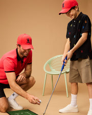 A man crouches while holding a golf ball, smiling and guiding a young boy who is preparing to hit the ball with a golf club. Both wear red caps and casual attire, with the man in a Psycho Bunny MENS KAYDEN PIQUE POLO - B6K675C200. The boy smiles in anticipation. A green chair is placed in the background against a plain backdrop.