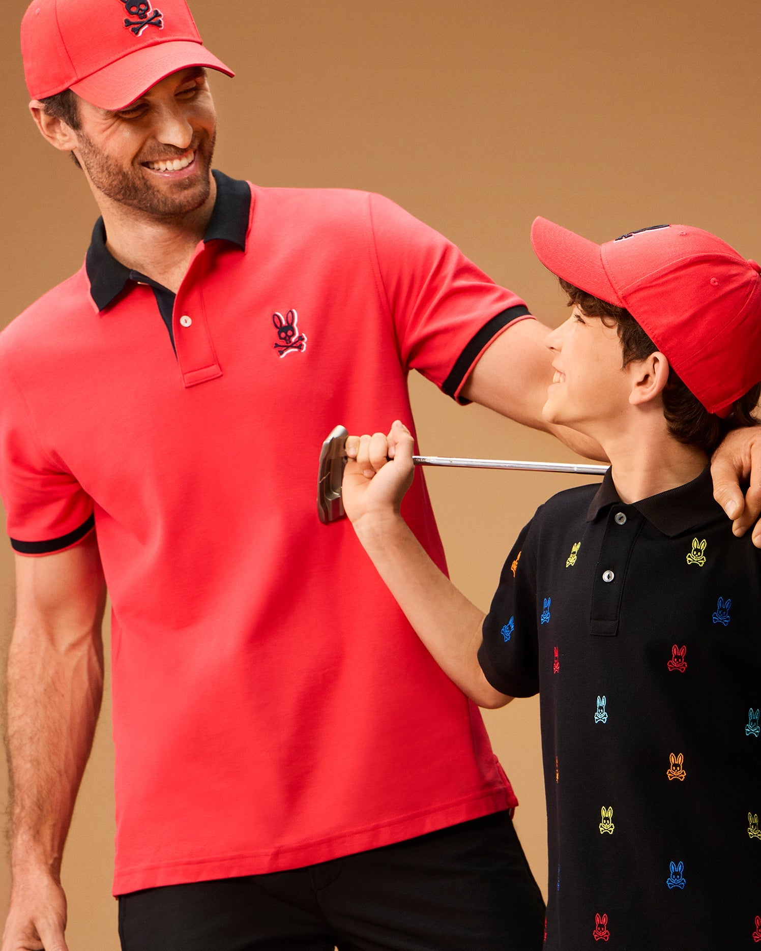 An adult and a child are smiling at each other. The adult is wearing a red cap and a red polo shirt with a rabbit logo, while the child is donning a Psycho Bunny MENS KAYDEN PIQUE POLO - B6K675C200 featuring colorful rabbit logos. The child, holding a golf club, sports contrasting stripes on their black cap.