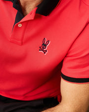 Close-up of a person wearing a red MENS KAYDEN PIQUE POLO - B6K675C200 from Psycho Bunny with black collar and sleeve trim. The shirt, made from diamond-knit Pima cotton, features an embroidered logo of a bunny with crossbones on the left chest. Only the upper torso and part of the arm are visible against a neutral background.