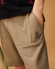 Close-up of a person wearing the Kids Stanford Light Pique Sweatshort in beige from Psycho Bunny, featuring a rabbit-shaped logo embroidered on the pocket. The person's hand is casually placed in the pocket, and they also wear a black shirt adorned with red rabbit-shaped logos. The background is a plain, light brown color—a stylish wardrobe staple.