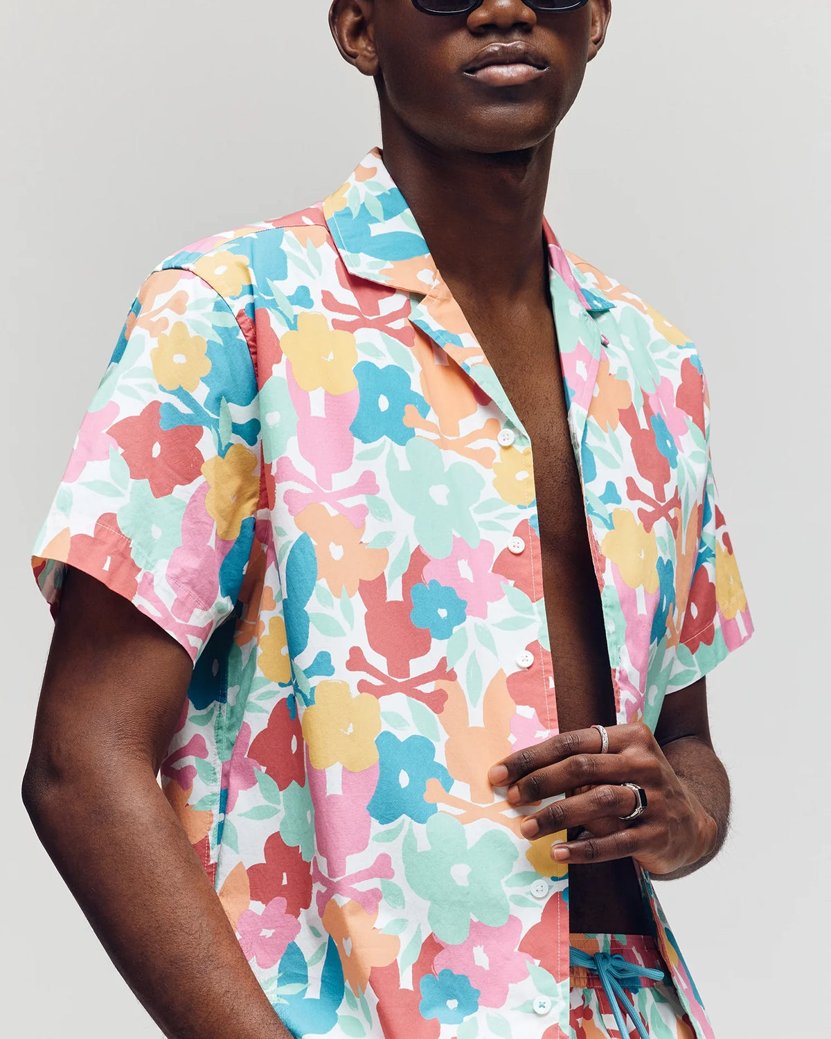 A person is wearing a vibrant Psycho Bunny MENS MEYLON STRETCH POPLIN SHIRT - MT0300034 with a camp collar, partially unbuttoned, paired with matching shorts. They sport sunglasses and adjust the shirt's edge with their left hand against a plain backdrop.