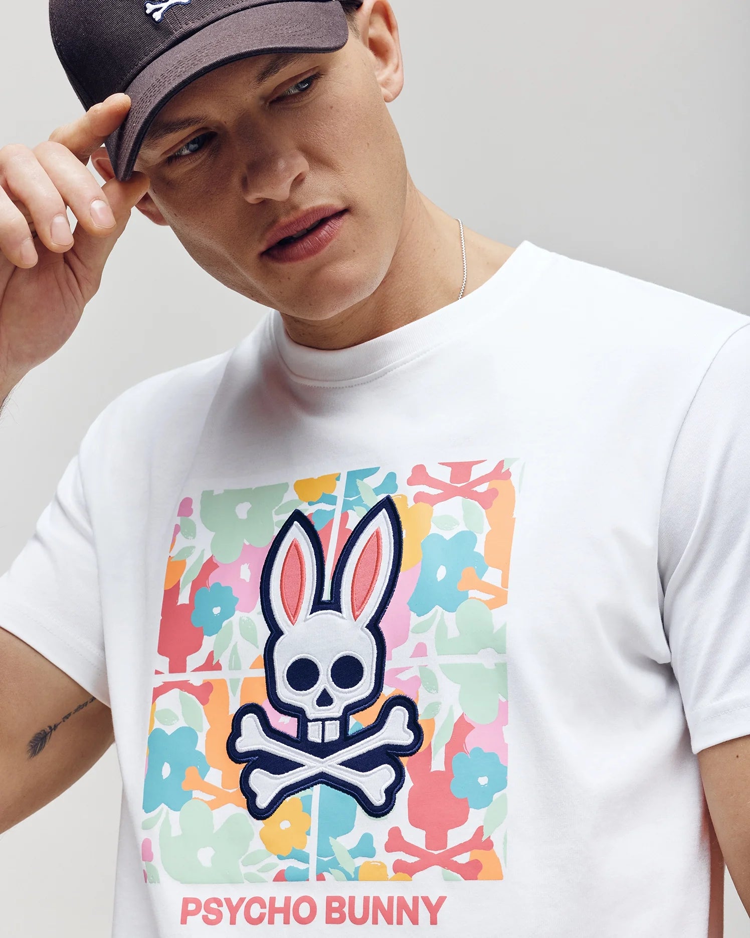 A person in a white Psycho Bunny MENS LEMORA GRAPHIC TEE - B6U657F200, featuring a colorful bunny skull and crossbones water-based print with "Psycho Bunny" at the bottom, adjusts their black cap against a plain backdrop.