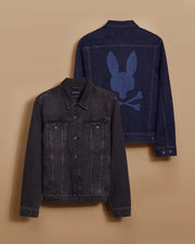 Displayed on a tan background are two men's denim jackets. The front jacket, labeled as the MENS PATHE DENIM JACKET - B6J741X1CO by Psycho Bunny, is black with buttons, multiple pockets, and an adjustable fit. The back jacket is dark blue and showcases embroidered details of a stylized skull and crossbones with bunny ears on the back.