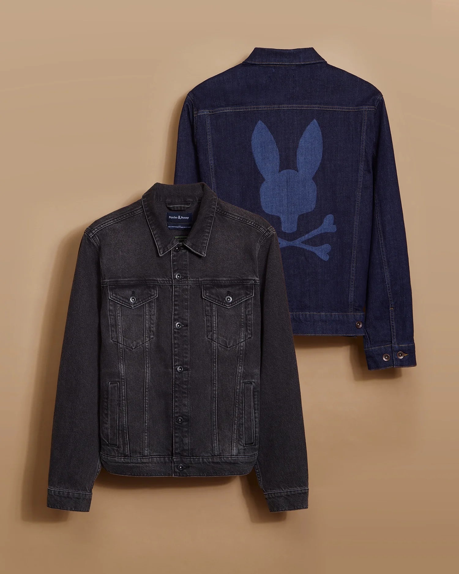 Displayed on a tan background are two men's denim jackets. The front jacket, labeled as the MENS PATHE DENIM JACKET - B6J741X1CO by Psycho Bunny, is black with buttons, multiple pockets, and an adjustable fit. The back jacket is dark blue and showcases embroidered details of a stylized skull and crossbones with bunny ears on the back.