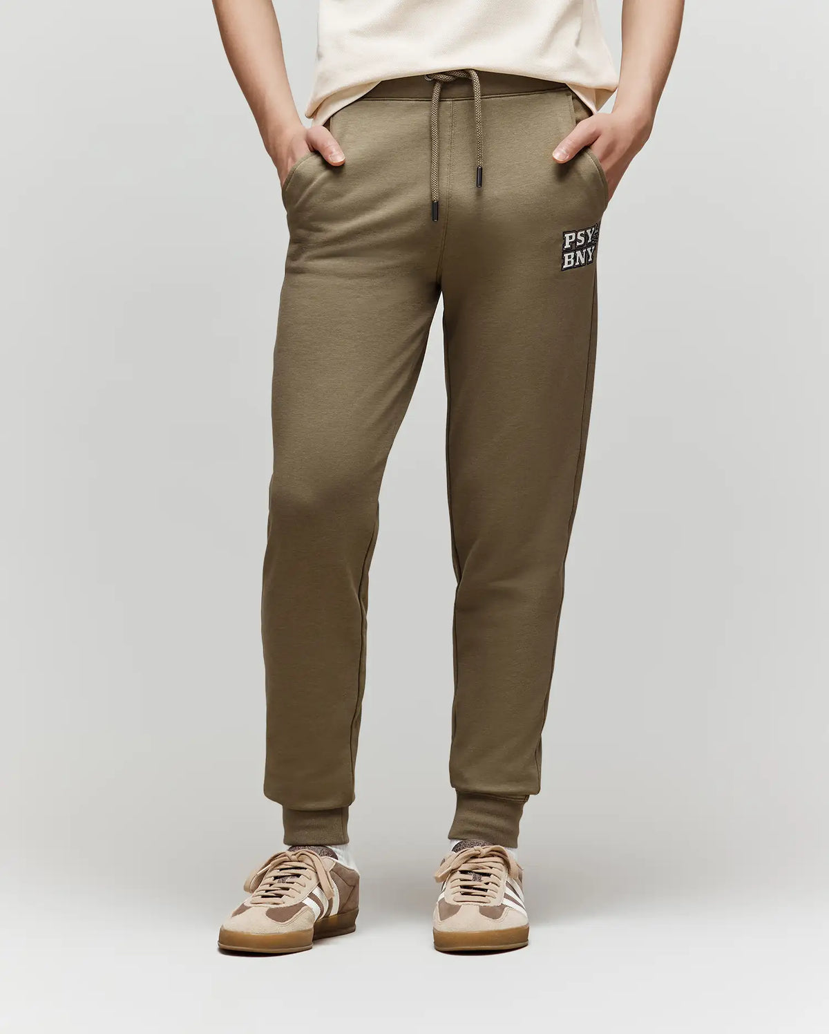 A child models the Psycho Bunny KIDS WADE SWEATPANT - KB0800002, an olive green jogger with a drawstring waist and white text on the left thigh, paired with a matching set, cream shirt, beige sneakers, hands in pockets. Background is plain light gray.