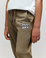 A child wearing Psycho Bunny KIDS WADE SWEATPANT - KB0800002 in olive green, featuring the "PSY BNY" logo and bunny graphic on the left thigh, made from a premium cotton blend, paired with a white t-shirt. Only torso and upper legs visible.