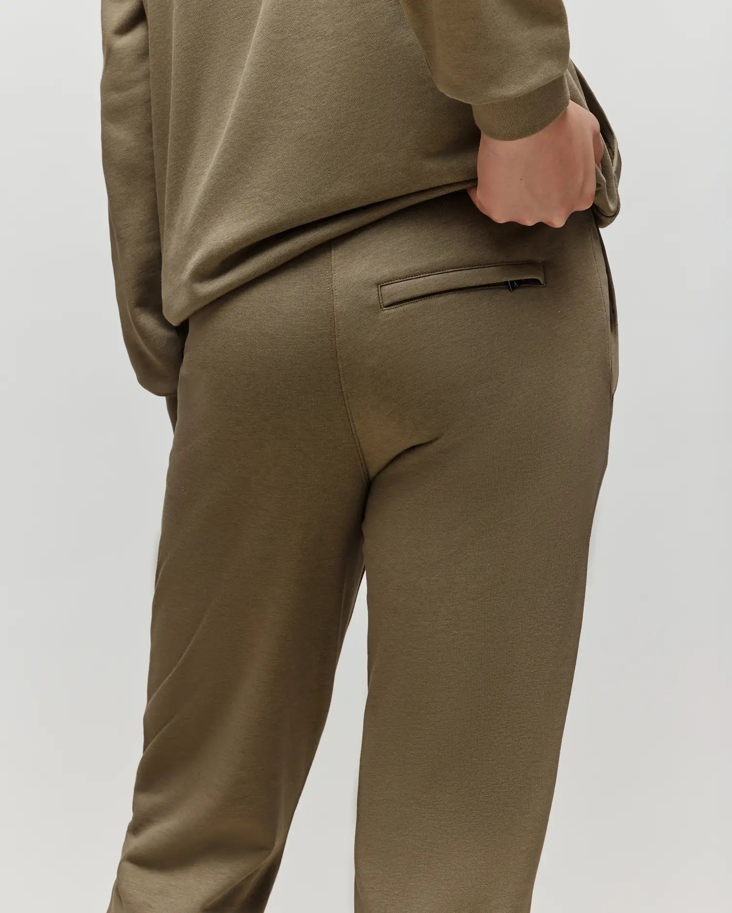 A child wears the Psycho Bunny KIDS WADE SWEATPANT (KB0800002) in olive green, premium cotton blend, posing from behind with one hand on the waistband near a welt pocket, set against a plain light background.