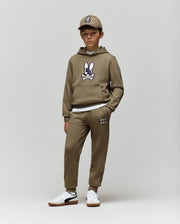A young boy faces forward against a plain background, wearing Psycho Bunny’s KIDS WADE SWEATPANT (KB0800002) in tan with jogger pants, a hoodie featuring the cartoon bunny logo, a matching cap, and white sneakers. His hands are in his pockets.