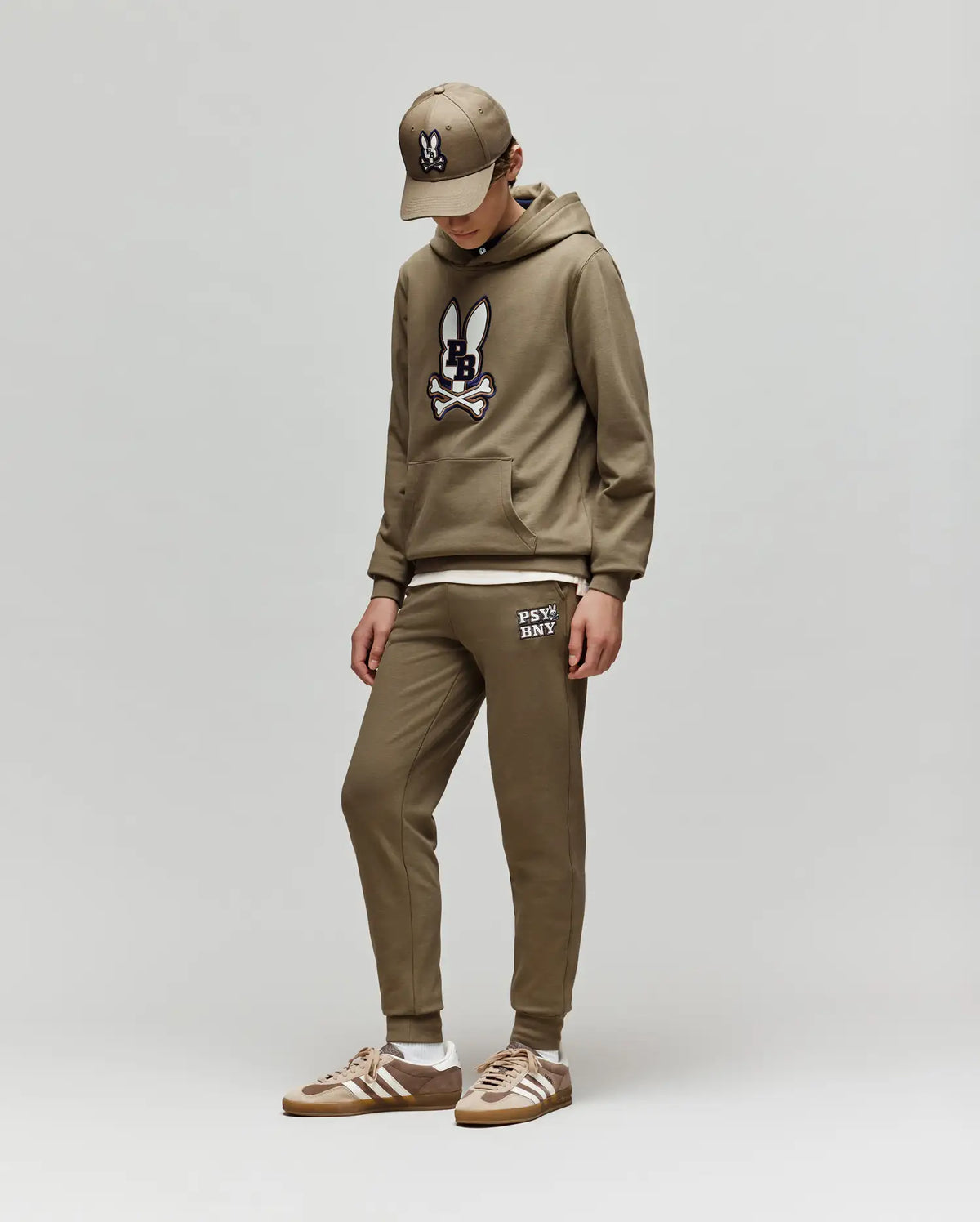 Wearing Psycho Bunny’s KIDS WADE SWEATPANT - KB0800002, a khaki hoodie with a bunny logo, cap, and brown Adidas sneakers, a person stands against a plain light gray background looking down.