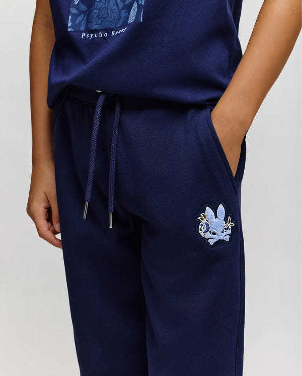 A person wearing Psycho Bunny KIDS AUGUSTE JOGGER - KB0800003 in 100% cotton navy blue with a light blue embroidered bunny skull logo and a matching navy shirt, standing with one hand in their pocket.