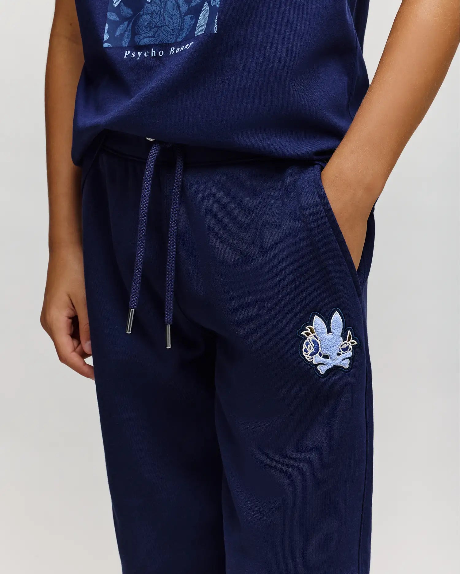 A person wearing Psycho Bunny KIDS AUGUSTE JOGGER - KB0800003 in 100% cotton navy blue with a light blue embroidered bunny skull logo and a matching navy shirt, standing with one hand in their pocket.