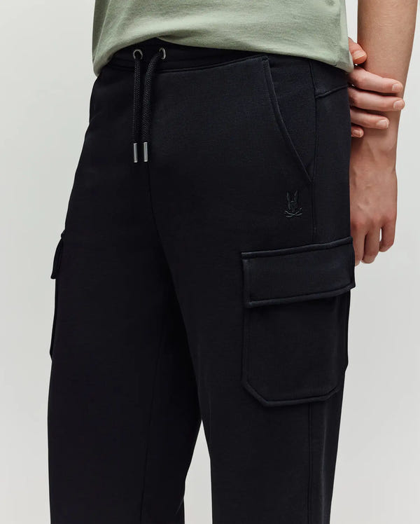 A person wearing Psycho Bunny KIDS NASH CARGO SWEATPANT - KB0800004—black cargo jogger sweatpants with large pockets, a drawstring waist, subtle stitching, and a small embroidered logo near the pocket—shown from the waist down.