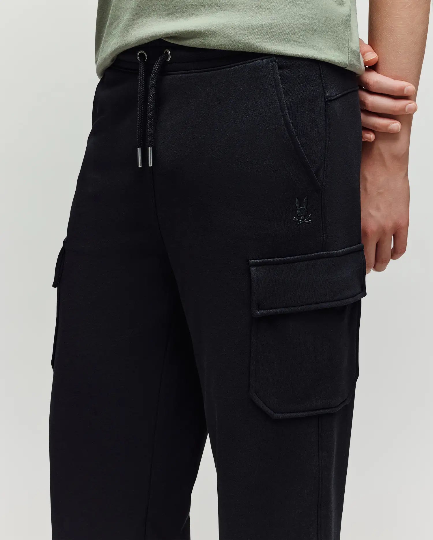 A person wearing Psycho Bunny KIDS NASH CARGO SWEATPANT - KB0800004—black cargo jogger sweatpants with large pockets, a drawstring waist, subtle stitching, and a small embroidered logo near the pocket—shown from the waist down.