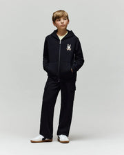 A young boy stands with hands in his pockets, wearing a black zip-up hoodie with Psycho Bunny’s cartoon bunny logo, KIDS NASH CARGO SWEATPANT (KB0800004) with cargo pockets, and white sneakers against a plain light gray background.