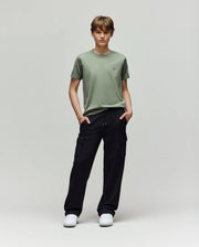 A young person stands in a studio against a light background, wearing Psycho Bunny KIDS NASH CARGO SWEATPANT - KB0800004 with handy cargo pockets, a light green tee, and white sneakers for ultimate sweatpant comfort. Hands are tucked in their pockets.