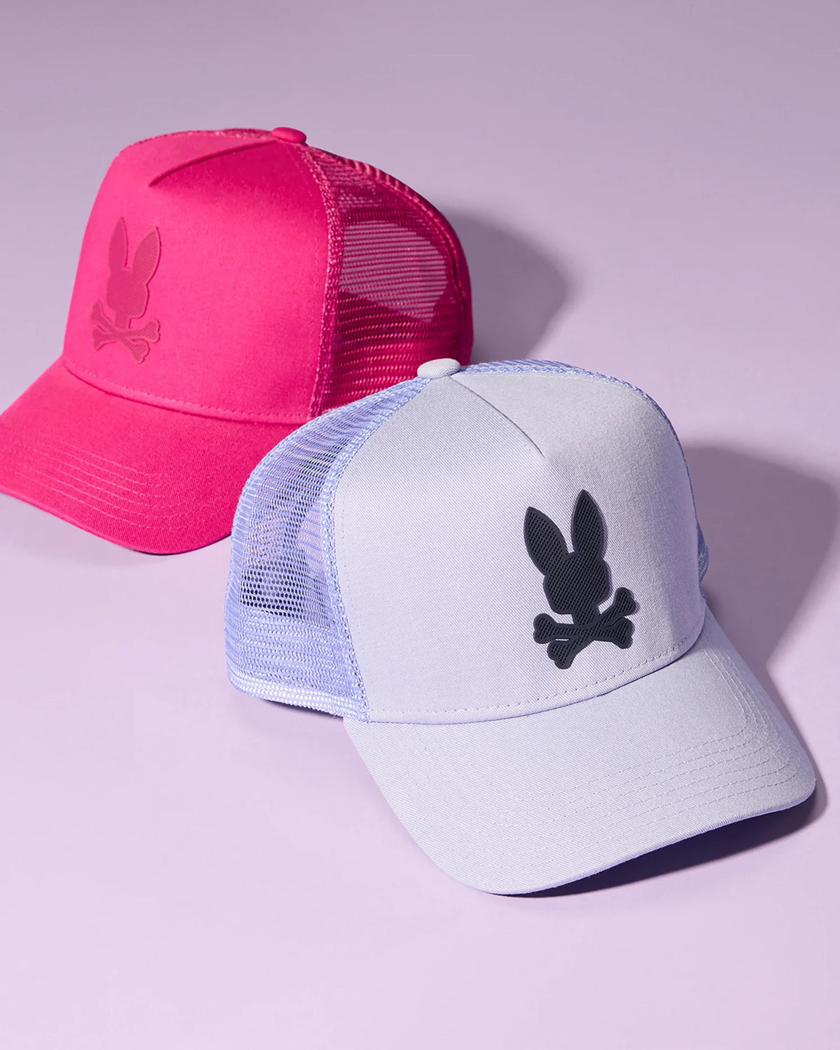 Two MENS RIVIERA TRUCKER CAPS - B6A667C200 by Psycho Bunny are displayed on a purple background. The cap in the foreground is light purple with a black bunny and crossbones logo, while the one behind it is bright pink with a matching bunny and crossbones logo. Both give off early 2000s nostalgia with their mesh back panels and snapback fastening.