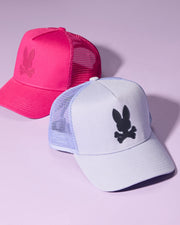 Two MENS RIVIERA TRUCKER CAPS - B6A667C200 by Psycho Bunny are displayed on a purple background. The cap in the foreground is light purple with a black bunny and crossbones logo, while the one behind it is bright pink with a matching bunny and crossbones logo. Both give off early 2000s nostalgia with their mesh back panels and snapback fastening.