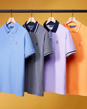 Four men's MENS WINTON PIQUE POLO SHIRT - B6K841A2PC by Psycho Bunny are hanging on wooden hangers against a yellow background. The shirts, crafted from luxurious Peruvian Pima cotton in a diamond-knit piqué, come in light blue, gray, lavender, and orange. Each features a small emblem on the left chest and a dark-colored collar.