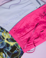 Three pairs of swim trunks are displayed on a light pink background. The top pair is light blue with "PSYCHO BUNNY" written in white, from the Psycho Bunny MENS MELVIN SWIM TRUNK collection (B6W665C200). The middle pair is vibrant pink with bunny designs, while the bottom pair features an abstract yellow, green, and blue pattern with a small bunny logo.