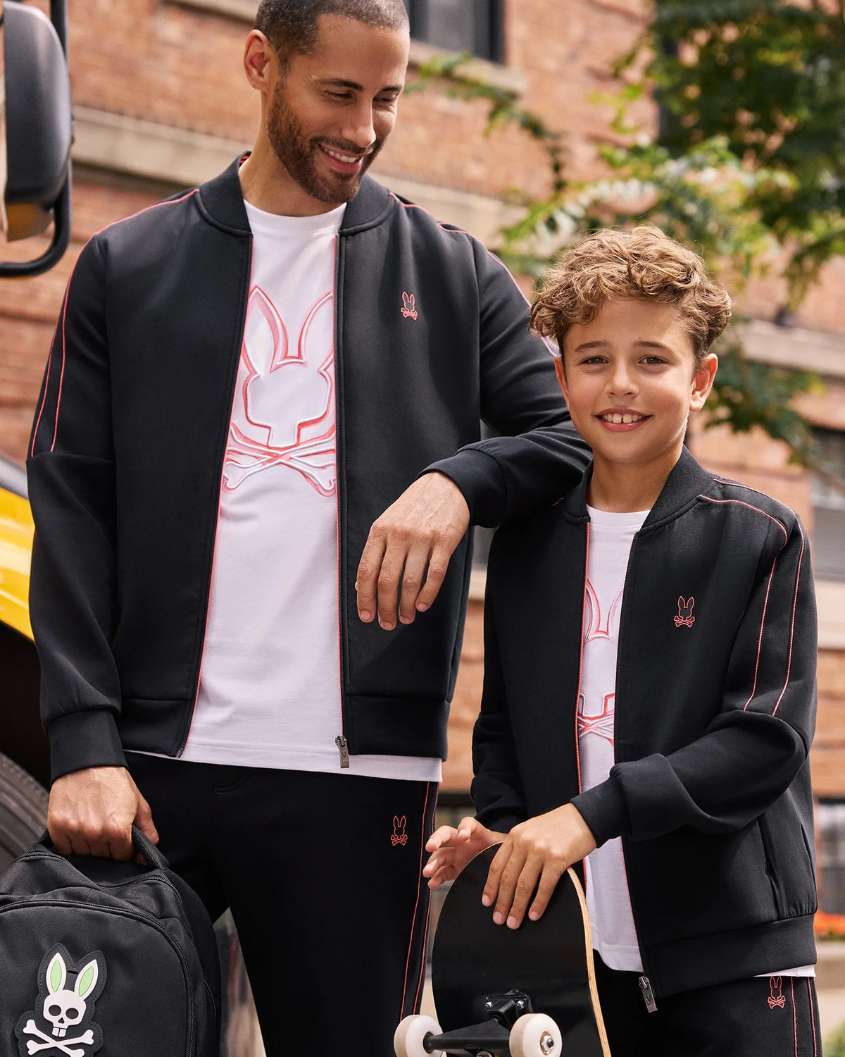 A man and a boy are smiling and wearing matching black jackets with white shirts featuring a neon bunny logo. The boy, dressed in KIDS TRAVIS TRACK PANT - B0P143D200 by Psycho Bunny, holds a skateboard while the man rests his hand on the boy's shoulder, holding a black backpack adorned with the same bunny logo. They stand outdoors.