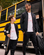 A man and a boy, both sporting matching black tracksuits and white t-shirts featuring a bunny logo, stand beside a yellow school bus. The boy holds a skateboard and looks up at the man, who smiles back while holding a black backpack. Their look is completed by the Psycho Bunny KIDS TRAVIS TRACK PANT - B0P143D200, which boasts elastic waistbands and contrast chainstitch stripes for added comfort and style.