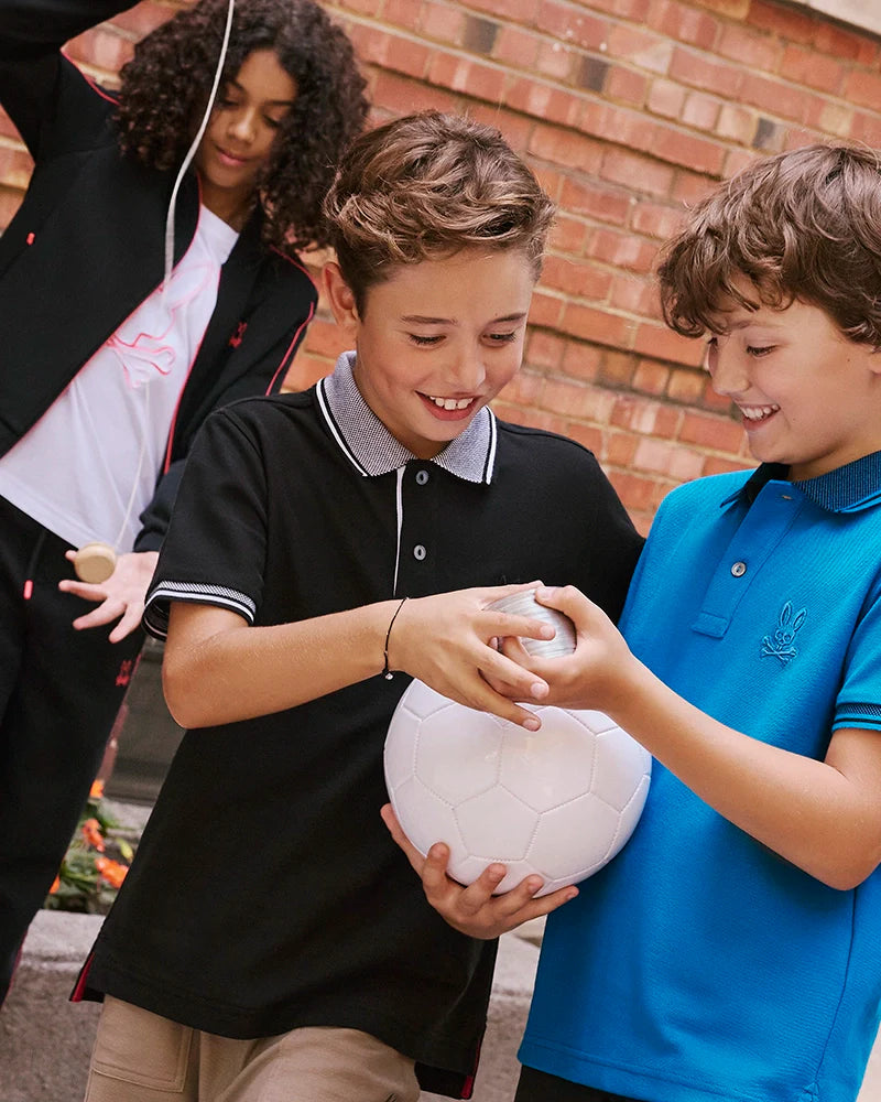 Three boys are gathered outdoors next to a brick wall. Two boys, dressed in Psycho Bunny's KIDS TRAVIS TRACK PANT - B0P143D200, which features elastic and comfortable design with contrast chainstitch stripes, are holding a soccer ball and smiling. Meanwhile, the third boy in the background is playing with a kendama toy. They appear engaged and happy, wearing casual clothes.