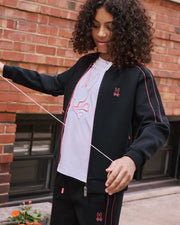 A person with curly hair is wearing a black jacket with pink piping and a bunny logo. They paired it with the KIDS TRAVIS TRACK PANT - B0P143D200 from Psycho Bunny, along with a white T-shirt featuring a larger bunny print. They stand by a brick wall holding a string or thread, while flowers in a pot are visible on the ground.