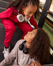 Two children with curly hair share a joyful moment on a bus. The one in red athletic wear and white sneakers sits on the steps, while the other, wearing a gray hoodie and Psycho Bunny's KIDS WALTER LIGHTWEIGHT SWEATPANT - B0P860D200 for a breathable fit, looks up at them, both smiling warmly at each other.