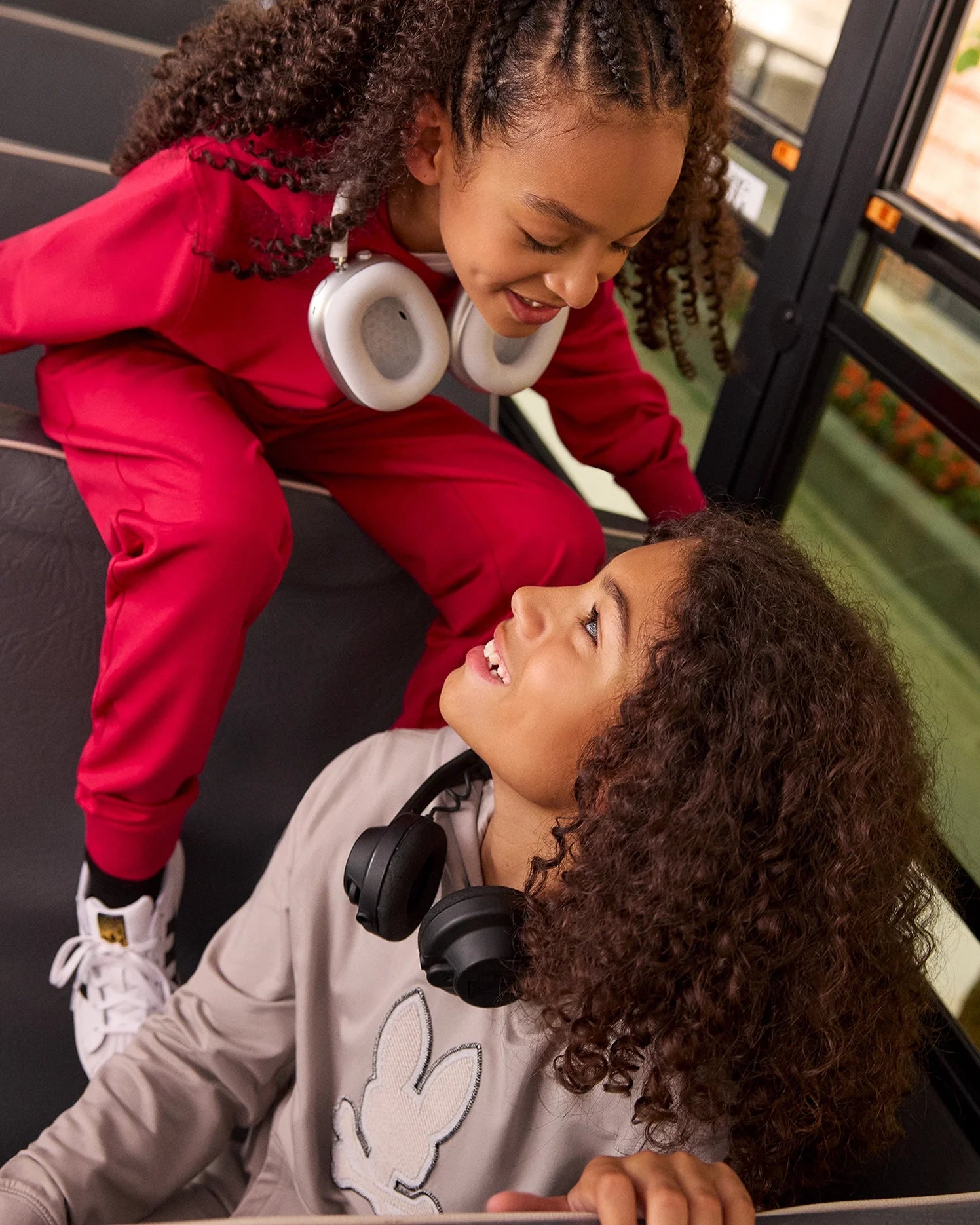 Two children with curly hair sit on a bus, smiling at each other. One child, wearing a red tracksuit with white headphones, is on a higher step. The other child, in Psycho Bunny's KIDS WALTER LIGHTWEIGHT HOODIE - B0H476D200 in beige and black headphones, looks up from a lower seat, giving the scene a modern twist.
