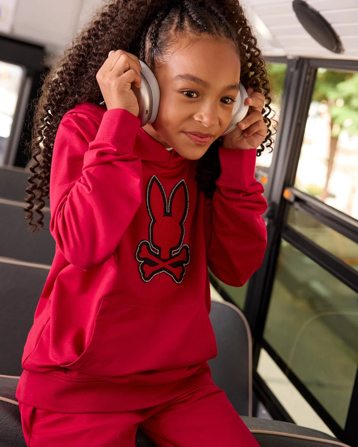 A young girl with curly hair sits on a bus, looking content as she listens to something through her silver headphones. Wearing a Psycho Bunny bright red KIDS WALTER LIGHTWEIGHT HOODIE (B0H476D200) featuring a rabbit and crossbones design, this modern twist crafted from soft Micro French terry is both stylish and comfortable. The bus window shows a blur of greenery outside.