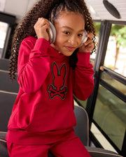 A young girl with curly hair sits on a bus, looking content as she listens to something through her silver headphones. Wearing a Psycho Bunny bright red KIDS WALTER LIGHTWEIGHT HOODIE (B0H476D200) featuring a rabbit and crossbones design, this modern twist crafted from soft Micro French terry is both stylish and comfortable. The bus window shows a blur of greenery outside.