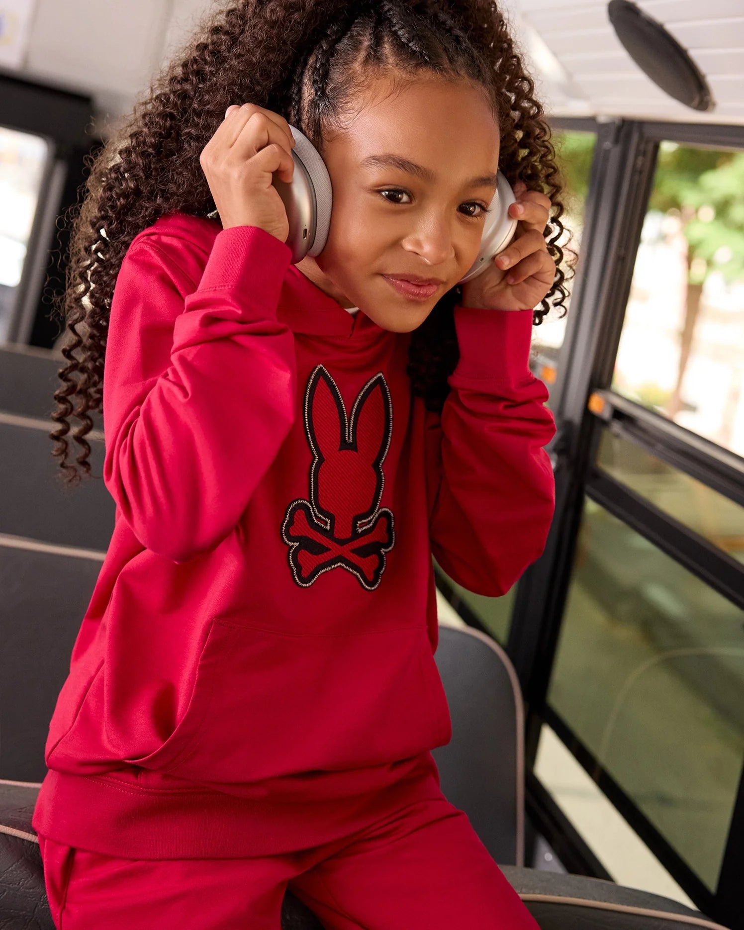 A young girl with curly hair sits on a bus, looking content as she listens to something through her silver headphones. Wearing a Psycho Bunny bright red KIDS WALTER LIGHTWEIGHT HOODIE (B0H476D200) featuring a rabbit and crossbones design, this modern twist crafted from soft Micro French terry is both stylish and comfortable. The bus window shows a blur of greenery outside.