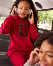 Two children are on a bus. The child in the foreground wears a grey Micro French terry hoodie and smiles while looking forward. The child in the background, sporting the red KIDS WALTER LIGHTWEIGHT HOODIE - B0H476D200 from Psycho Bunny, brings a modern twist by listening to headphones and glancing to the side.